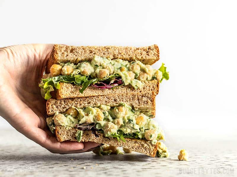 Eine Hand greift nach einem Stapel aus zwei Hälften eines Sandwiches mit Frühlingszwiebel- und Kräuter-Kichererbsensalat auf Weizenbrot.