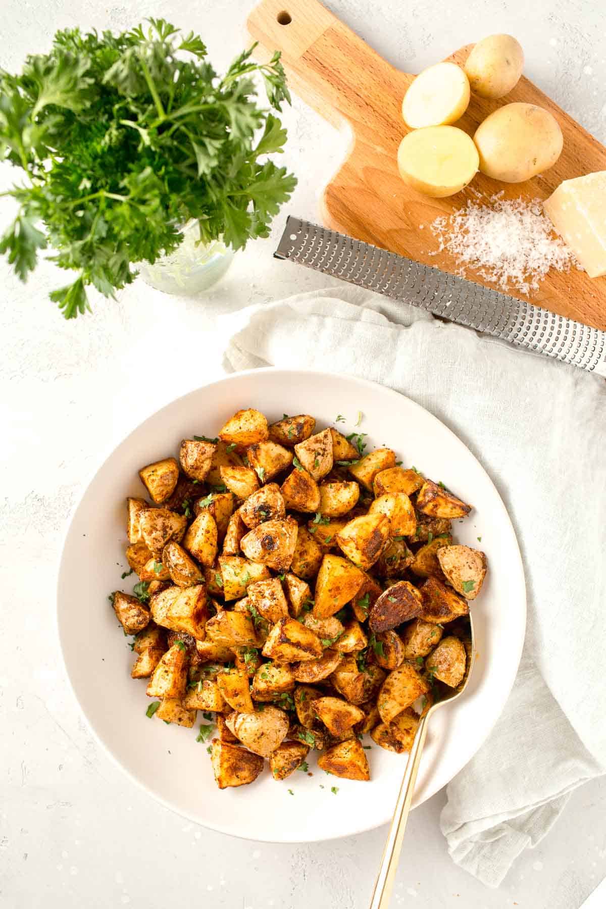 Overhead-Bild von gerösteten Parmesankartoffeln in einer großen weißen Schüssel neben einem Leinentuch, frischer Petersilie, einer Metallreibe und frisch geriebenem Parmesankäse