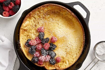 Overhead view of a dutch baby in a cast iron skillet with fresh berries in the center and maple syrup on the side.